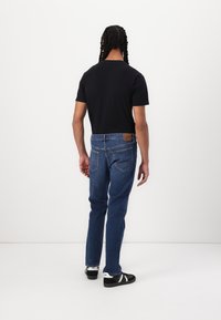 Mørk t-skjorte og blå jeans sett bakfra. Jeansene har en brun lærpatch med "Lee"-merke og subtile sømdetaljer.