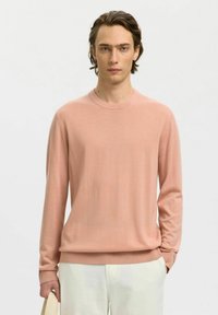 Jeune homme portant un pull léger à manches longues rose pâle et un pantalon écru, tenant un sac fourre-tout beige dans sa main gauche.