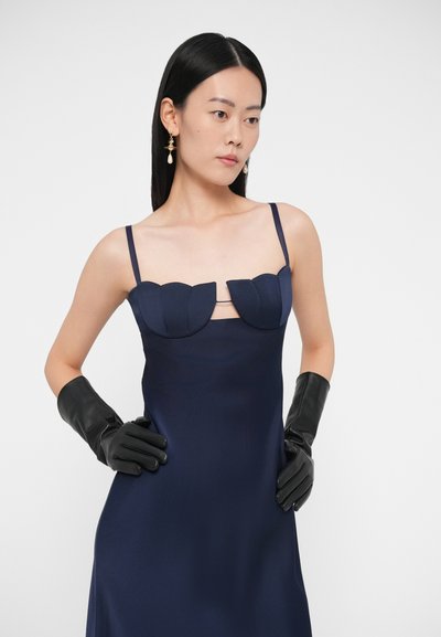 Sieviete ar taisniem melniem matiem valkā tumši zilu bezroku kleitu ar viļņotu izgriezumu un melnas ādas cimdus, pozējot ar rokām uz gurniem.