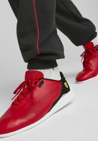 Czerwone skórzane sneakersy z perforowanymi detalami, czarnymi akcentami i białą podeszwą. W połączeniu z szarymi spodniami i białymi skarpetkami.