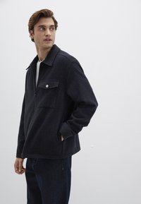 Marineblaues Zip-Up-Jackett aus strukturiertem Material, mit spitzem Kragen, einer Vorderasche mit silbernem Knopf und lockerer Passform. Zutragen mit dunklen Jeans.