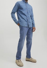 Camisa de mezclilla azul claro con botones y un bolsillo en el pecho, combinada con jeans de mezclilla azul más claro y zapatos de ante beige, presentada en un modelo.