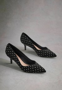 Svart nubuck högklackade pumps med spetsiga tår, prydda med vita pärlliknande dekorationer, har en elegant, slät insida.