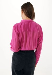 Camisa de satén rosa con mangas largas, cuello y detalle de pliegues en la parte posterior, combinada con jeans negros de cintura alta. Textura suave, color vibrante.