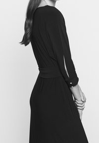 Žena v černých dlouhých šatech s rozparkem a knoflíky, s rukama sepjatýma, vlasy v volných vlnách, na jednobarevném pozadí.