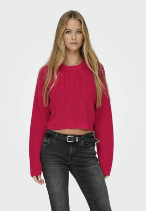 Un pull court rose vif avec un motif tricoté texturé, doté de longues manches et d'un col rond, associé à un jean noir et une ceinture.