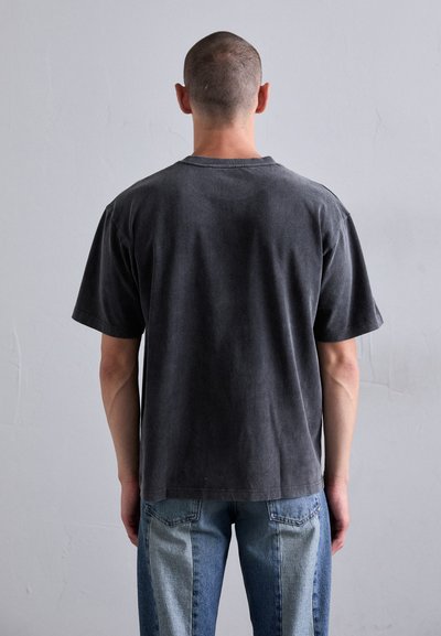 Mann mit kurzem Haar, der ein schwarzes T-Shirt und zweifarbige blaue Jeans trägt, steht vor einer schlichten hellgrauen Wand.