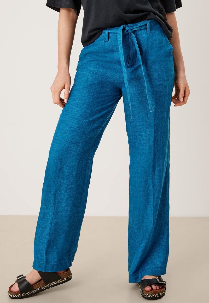 Pantalon ample en lin bleu léger avec taille à cordon. Texture lisse et coupe décontractée, associé à des sandales noires avec une bride et une semelle tissée.