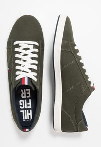 Gröna canvas sneakers med vita snören, kontrasterande marinblå inuti, gummisula och en liten röd, vit och blå logotyp vid hälen.