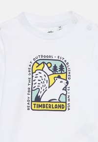 Bela bomba iz 100% bombaža s barvno ilustrirano logotipom veverice, gora in dreves, z napisom "TIMBERLAND" ter frazami, povezanimi z naravo.