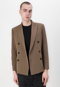 Blazer de doble botonadura en tejido marrón, con botones negros, solapas anguladas y un bolsillo en el pecho. Llevado sobre una camisa negra.