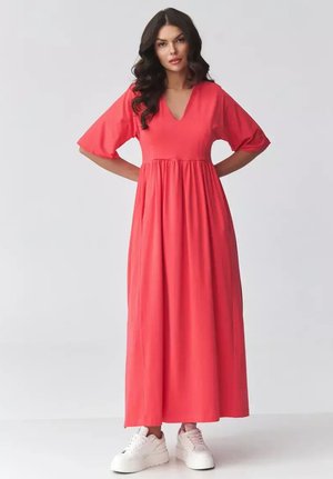 Kobieta o długich ciemnych włosach, ubrana w koralowo-czerwoną maxi sukienkę z dekoltem w serek oraz białe sneakersy na koturnie, stojąca z rękami na biodrach.