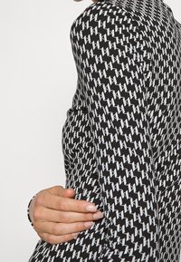 Maglia a maniche lunghe nera con un motivo geometrico bianco. Il materiale appare liscio con un design aderente. Presenta un orlo curvo e dettagli ai polsi.