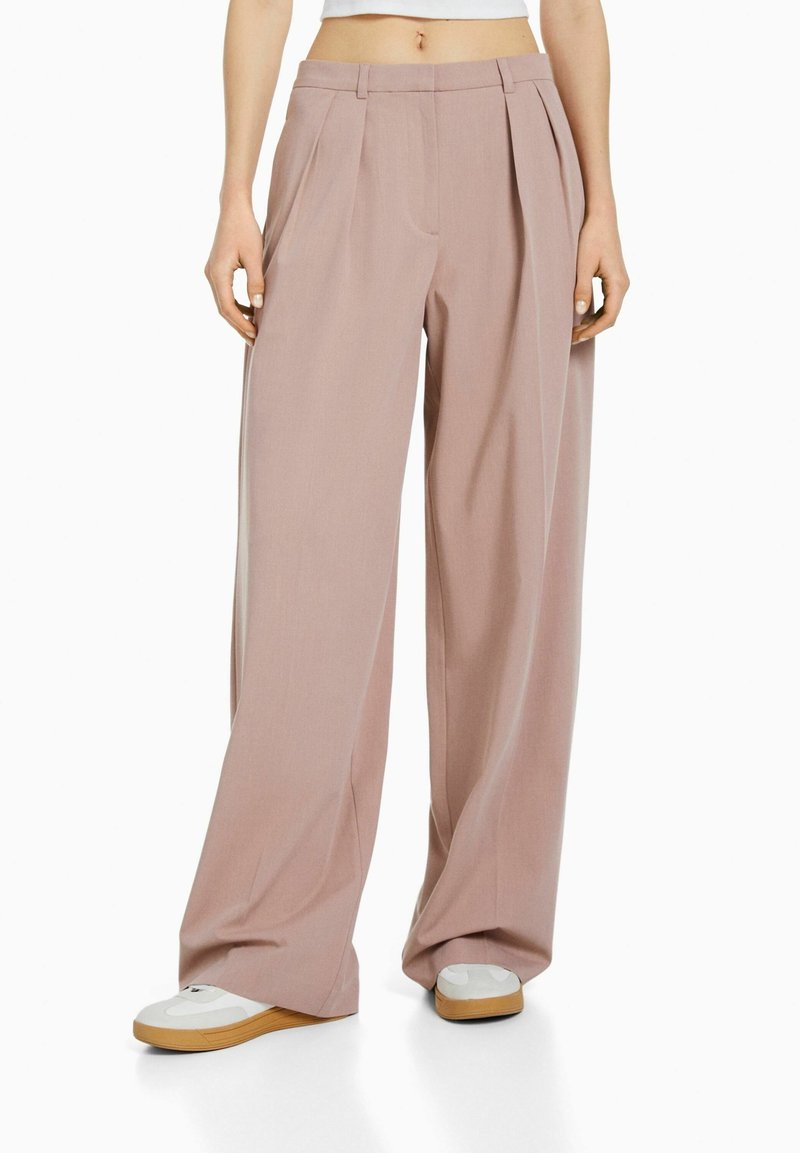 Videbeniga byxor i mjuk rosa tyg, med plisseringar, skräddarsydd midja och en slät yta, kombinerat med vita och bruna skor.