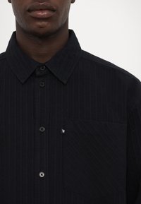 Svart collared skjorte laget av teksturert stoff, med vertikale striper, frontknapper, og en enkelt lomme med en logotag.