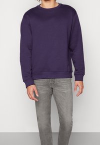 Sudadera morada con cuello redondo, puños y dobladillo acanalados, combinada con jeans gris claro. Textura suave, ajuste relajado, diseño minimalista.