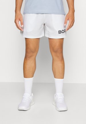Homme portant un short de sport blanc avec des lettres noires, des chaussettes blanches et des chaussures de sport blanches, debout devant un fond uni.