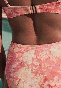 Ziedu bikini tops rozā un persiku nokrāsās ar teksturētu audumu. Iezīmē metāla aizdare un ziedu etiķete uz jostas.