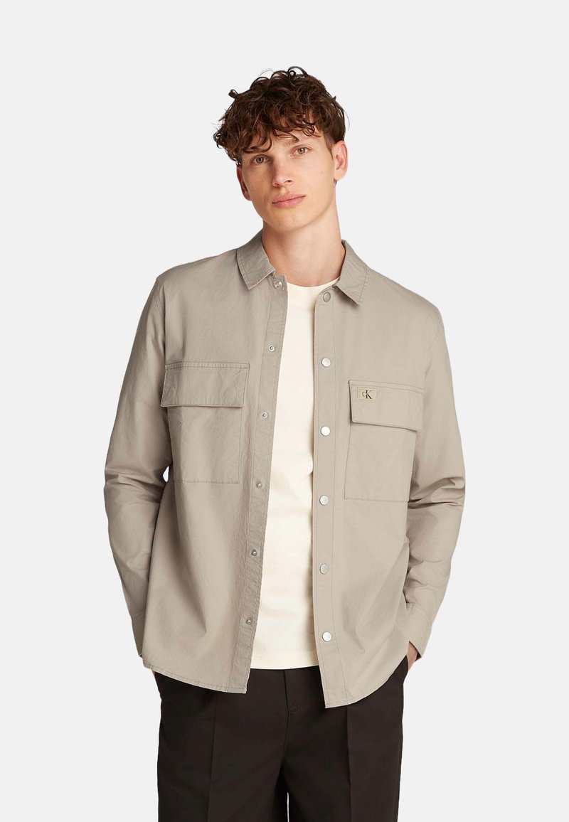 Camisa de botón color beige claro con dos bolsillos en el pecho, hecha de una tela texturizada. Presenta un cuello y botones a presión plateados, superpuesta sobre una blusa crema.