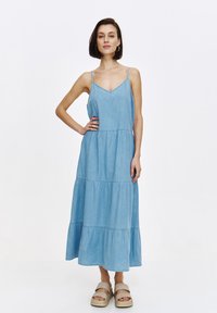 Hellblaues Denim-Maxi-Kleid mit dünnen Spaghetti-Trägern, einem V-Ausschnitt und einem gestuften Rockdesign. Modell trägt beigefarbene Plateausandalen.