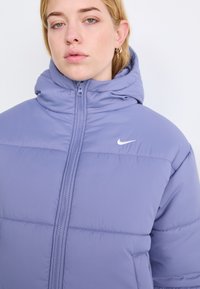 Svetlo vijolična puhovka s visokim kapuco, z zadrgo in majhnim belim logotipom Nike na prsih. Ima prešito zasnovo in gladko teksturo.