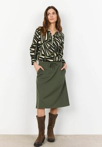 Chemise à manches longues à motifs en noir, vert et crème avec un col en V, associée à une jupe unie verte longueur genou et des bottes marron.