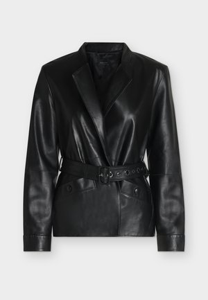 Femme aux longs cheveux foncés portant une veste et un pantalon en cuir noir, chemise blanche, tenant un petit sac à main argenté, debout sur fond blanc.