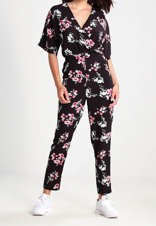 Jumpsuit noir à fleurs avec un décolleté en V, des manches courtes et un pantalon longueur cheville. Présente des motifs de fleurs roses et blanches, porté avec des baskets blanches.