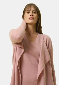 Ljusrosa kofta med V-ringning, gjord av mjukt tyg och har en öppen front samt draperad design. Slät textur, avslappnad stil.