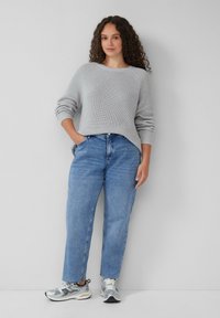 Hellgrauer Strickpullover mit Rundhalsausschnitt, kombiniert mit blauen, schmal geschnittenen Jeans und silbernen Sneakers. Strukturiertes Material und lässige Passform.