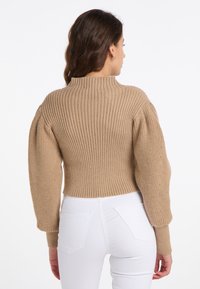 Maglione beige a costine con colletto alto e maniche lunghe a palloncino, abbinato a pantaloni bianchi aderenti. Texture a maglia visibile in tutto il capo.