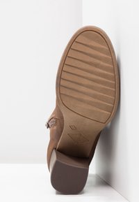 Brązowy zamszowy botek do kostki z teksturowaną gumową podeszwą, z lekkim obcasem i przeszyciem po boku. Płaska, żłobiona faktura podeszwy.