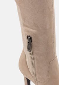 Stivale da donna in suede beige con cerniera laterale e tacco nero, mostrato in dettaglio ravvicinato su sfondo bianco.