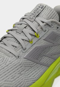 Κοντινό πλάνο γκρι αθλητικού παπουτσιού με κίτρινες λεπτομέρειες, περίτεχνες κορδόνια, υφή ύφασματος και ετικέτα «FuelCell Propel» στη γλώσσα.