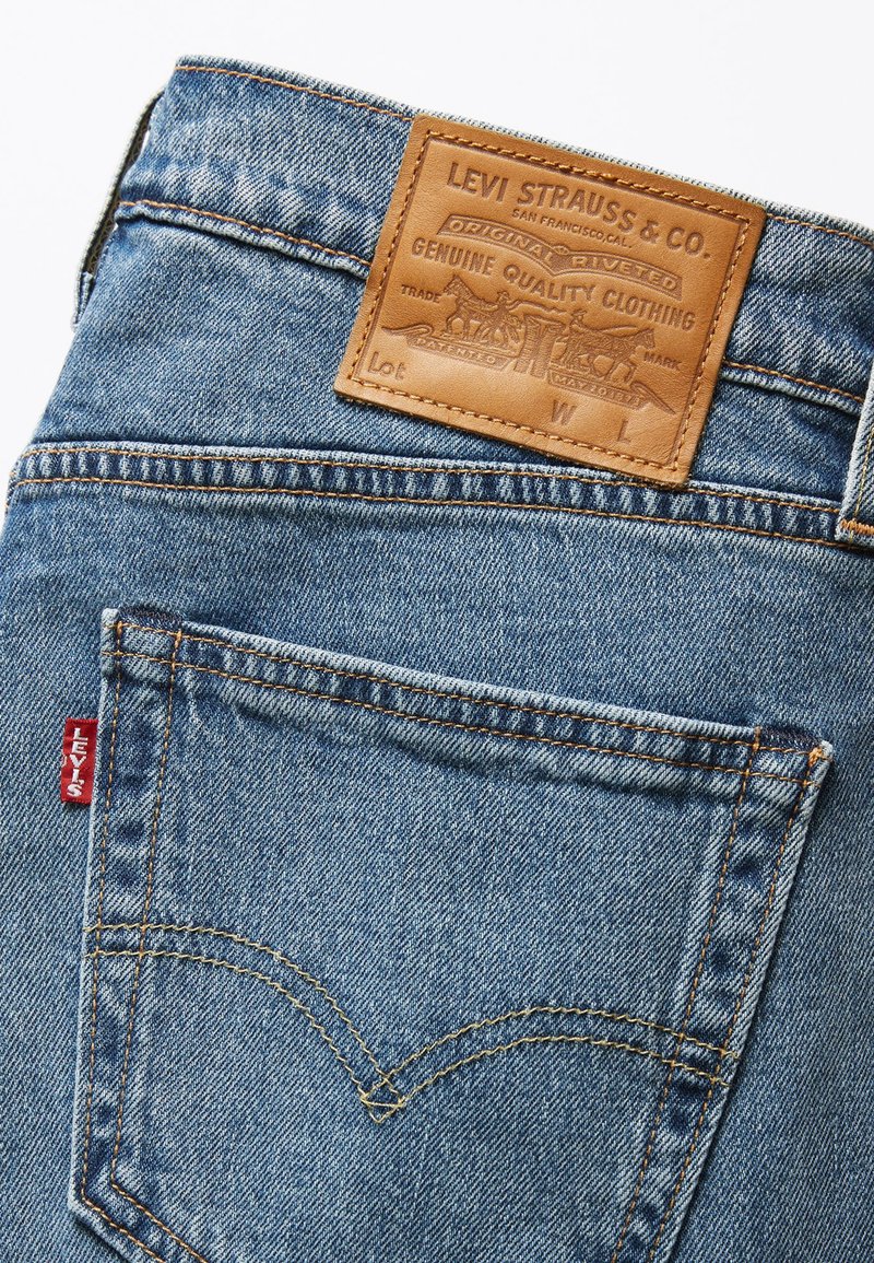 Jeans de mezclilla azul que presentan un parche de cuero con texto en relieve, una etiqueta de logo roja y un contraste de costuras amarillas en el bolsillo trasero.
