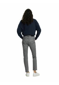 Personne aux longs cheveux bouclés, portant un pull bleu marine, un jean gris taille haute et des baskets blanches, debout les mains sur les hanches, de dos.