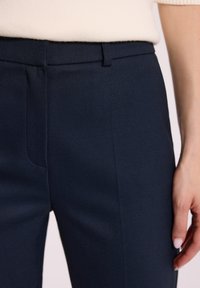 Zbliżenie na osobę w granatowych spodniakach szytych na miarę i jasnobeżowej bluzce, z ręką opartą obok spodni.