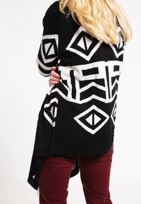 Cárdigan en blanco y negro con patrones geométricos, con un ajuste holgado y un dobladillo asimétrico. Combinado con pantalones burdeos.