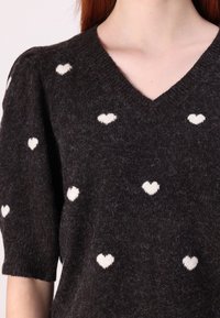 Pull noir en tricot avec manches courtes bouffantes, orné de motifs de cœurs blancs partout et un décolleté en V. Tissu doux et texturé.