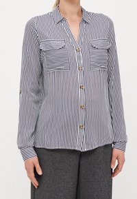 Camicia a righe con bottoni in verticale blu navy e bianco, dotata di due tasche sul petto e bottoni dorati. Maniche lunghe con dettagli sui polsini.