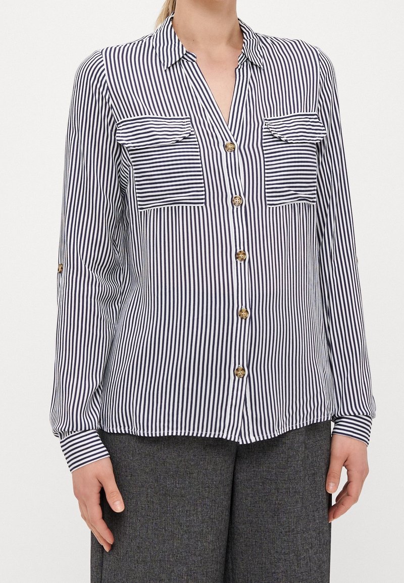 Camicia a righe con bottoni in verticale blu navy e bianco, dotata di due tasche sul petto e bottoni dorati. Maniche lunghe con dettagli sui polsini.