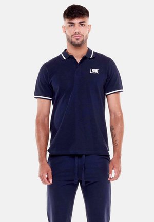 Uomo in piedi rivolto in avanti che indossa una polo blu navy con bordo bianco e logo, e pantaloni blu navy abbinati con coulisse.