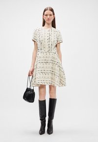 Kleita līdz ceļiem krēmkrāsā ar melniem rakstiem, ar īsām piedurknēm un izplūdušu svārku. Saskaņota ar melnām zābakiem līdz ceļiem un mazu somu.