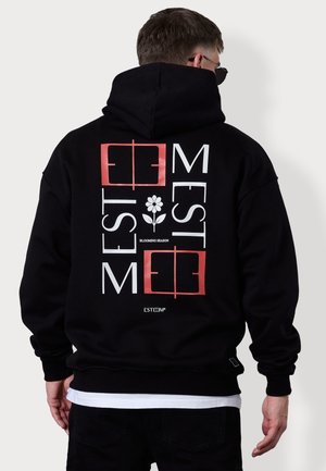 Mann trägt schwarzen Hoodie mit rotem und weißem Schriftzug "ESTM" und einer Blumenikone auf der Rückseite, steht vor weißem Hintergrund.