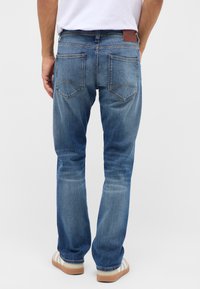Sinine denim koos sirge jala lõikega, tagakottide ja pruunist nahast plaadi detailiga vööl. Pleekinud viimistlus koos peente kulumise märkidega.