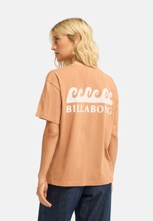 Blonde vrouw draagt een losse beige t-shirt met wit golfpatroon en de tekst "Billabong" op de achterkant, gecombineerd met donkerblauwe jeans.