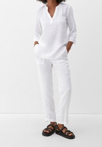 Weißes Leinenoberteil mit langen Ärmeln, V-Ausschnitt und Knopfleiste, kombiniert mit passenden, locker sitzenden Leinenhosen und schwarzen Riemchensandalen.