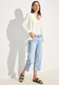 Hellgrüne Knopfhemd über einem weißen Tanktop, kombiniert mit hellblauen, weit geschnittenen Jeans und olivgrünen Sandalen mit Schnallen.