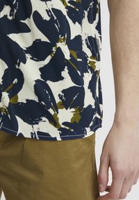 Camisa de manga corta en blanco con un patrón abstracto de pinceladas en azul marino y verde oliva. Tejido suave, bordes completados, etiqueta lateral visible.
