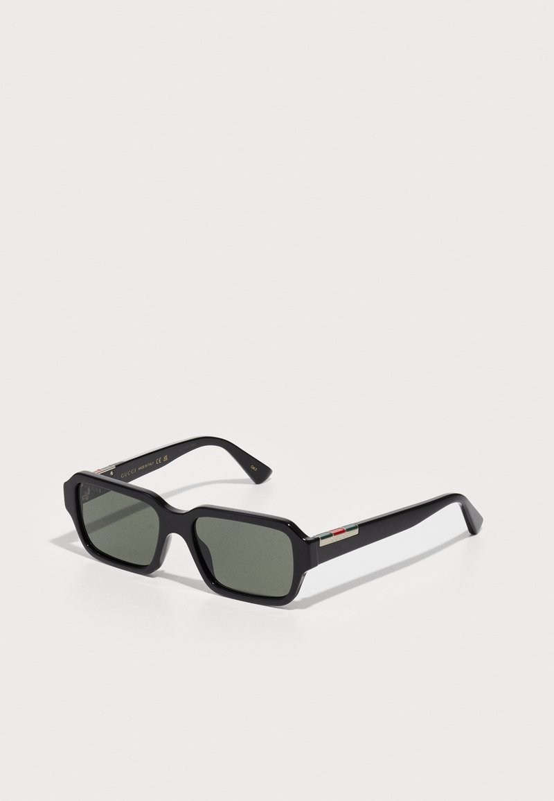 Lunettes de soleil noires rectangulaires avec des verres vert foncé, des montures épaisses et des accents matériels distinctifs en rouge, blanc et vert sur les branches.